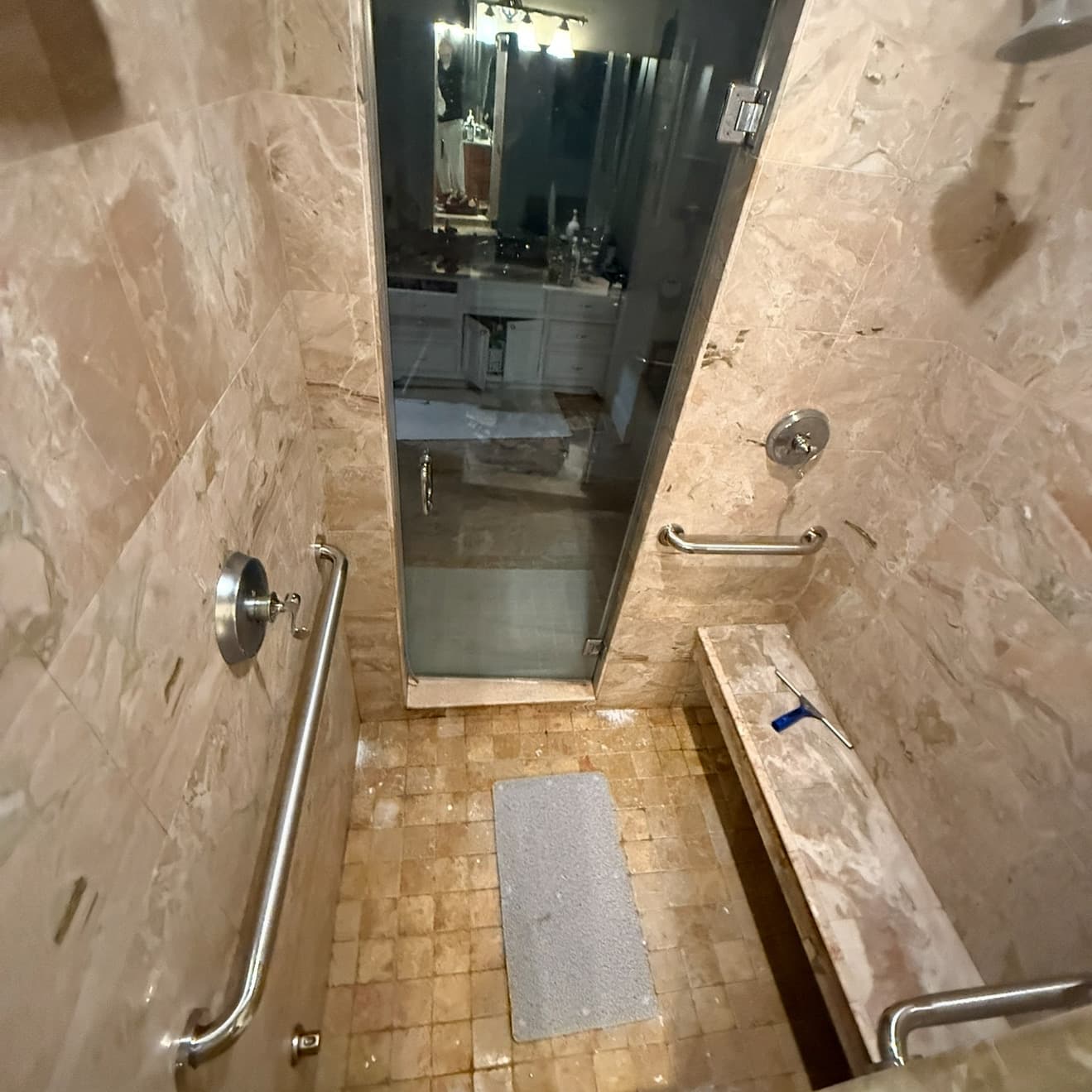 Tan marble walk-in shower featuring safety grab bars, a built-in bench, and a glass door.