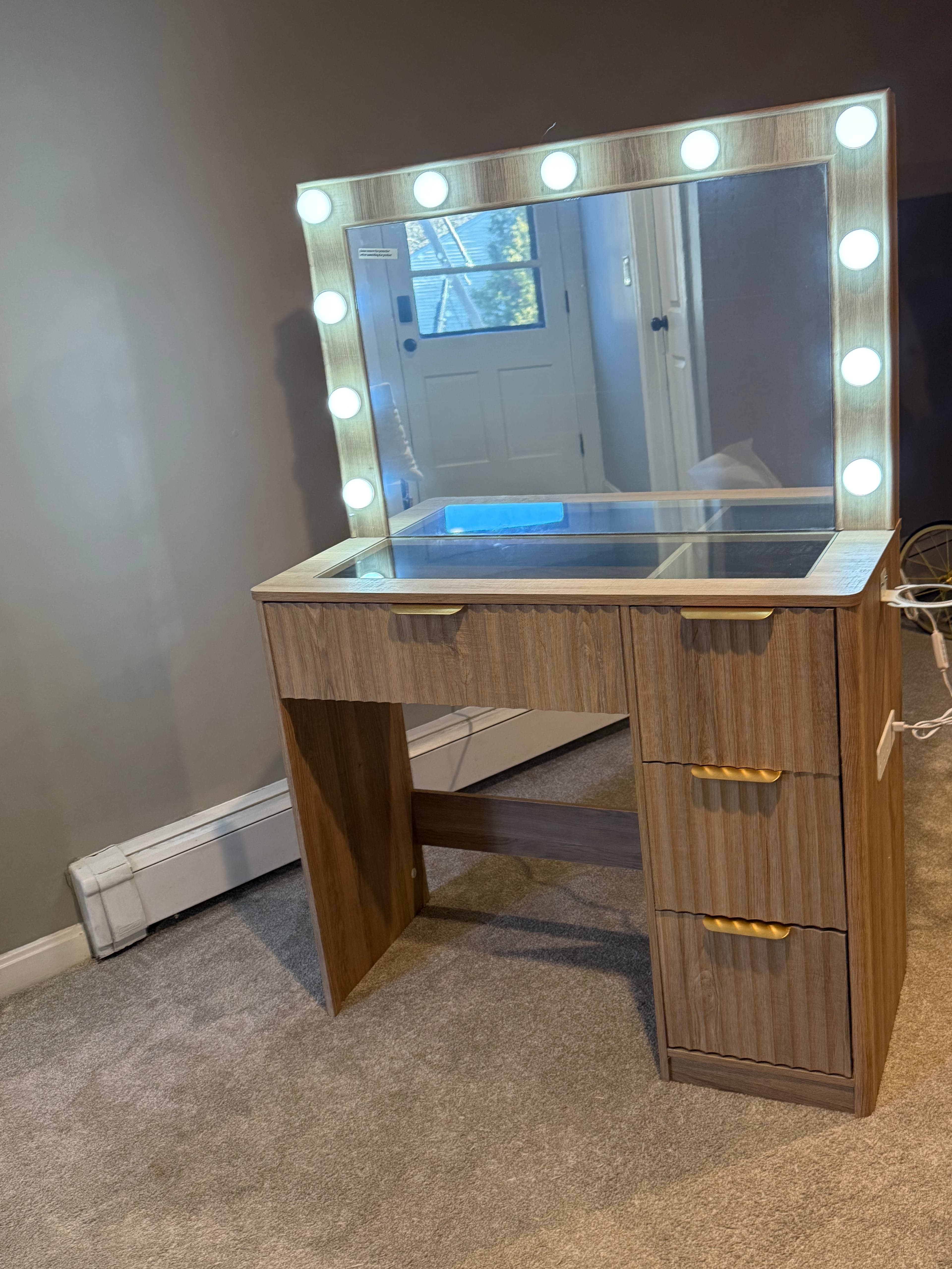 Wooden vanity desk with a Hollywood-style lighted mirror and drawers with gold handles.