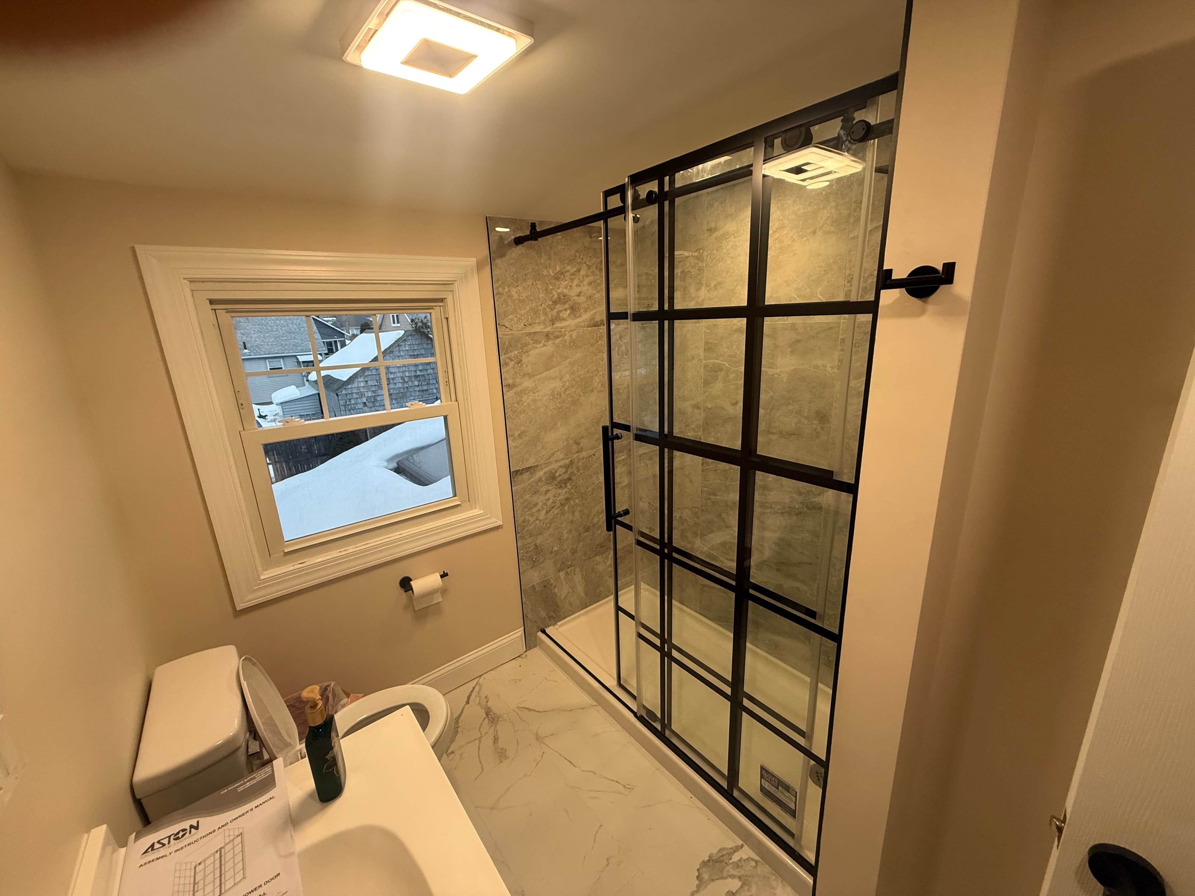 Modern bathroom with black grid shower door, marble tiling, and a window overlooking snow.