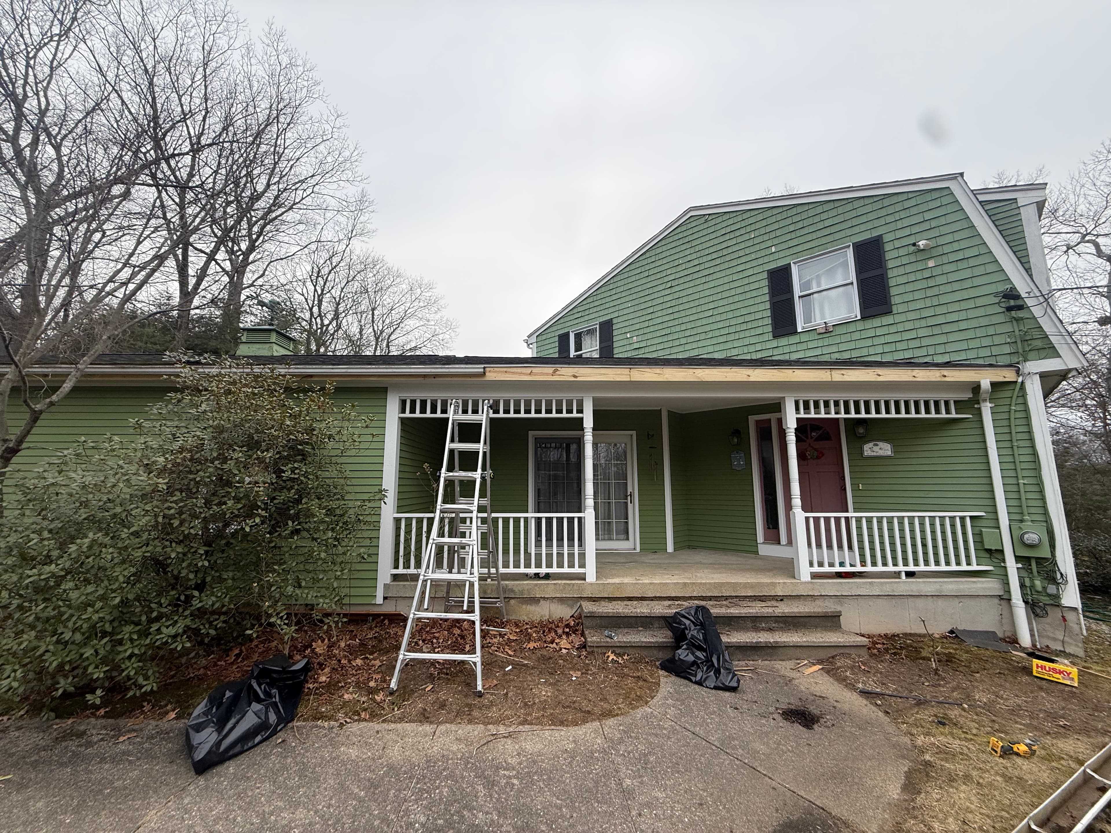 Silver ladder against a green house with a white porch undergoing roof trim repairs.