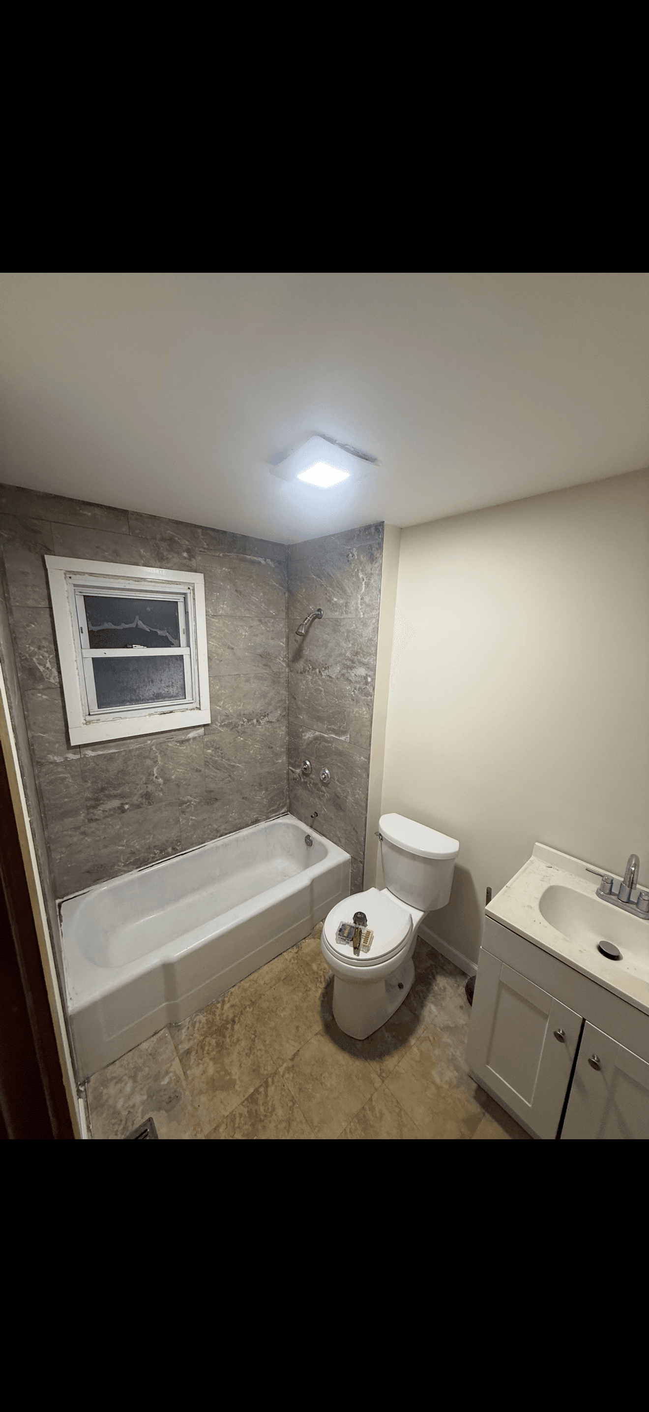 Bathroom with grey marble tile shower, white bathtub, toilet, and vanity sink under bright light.