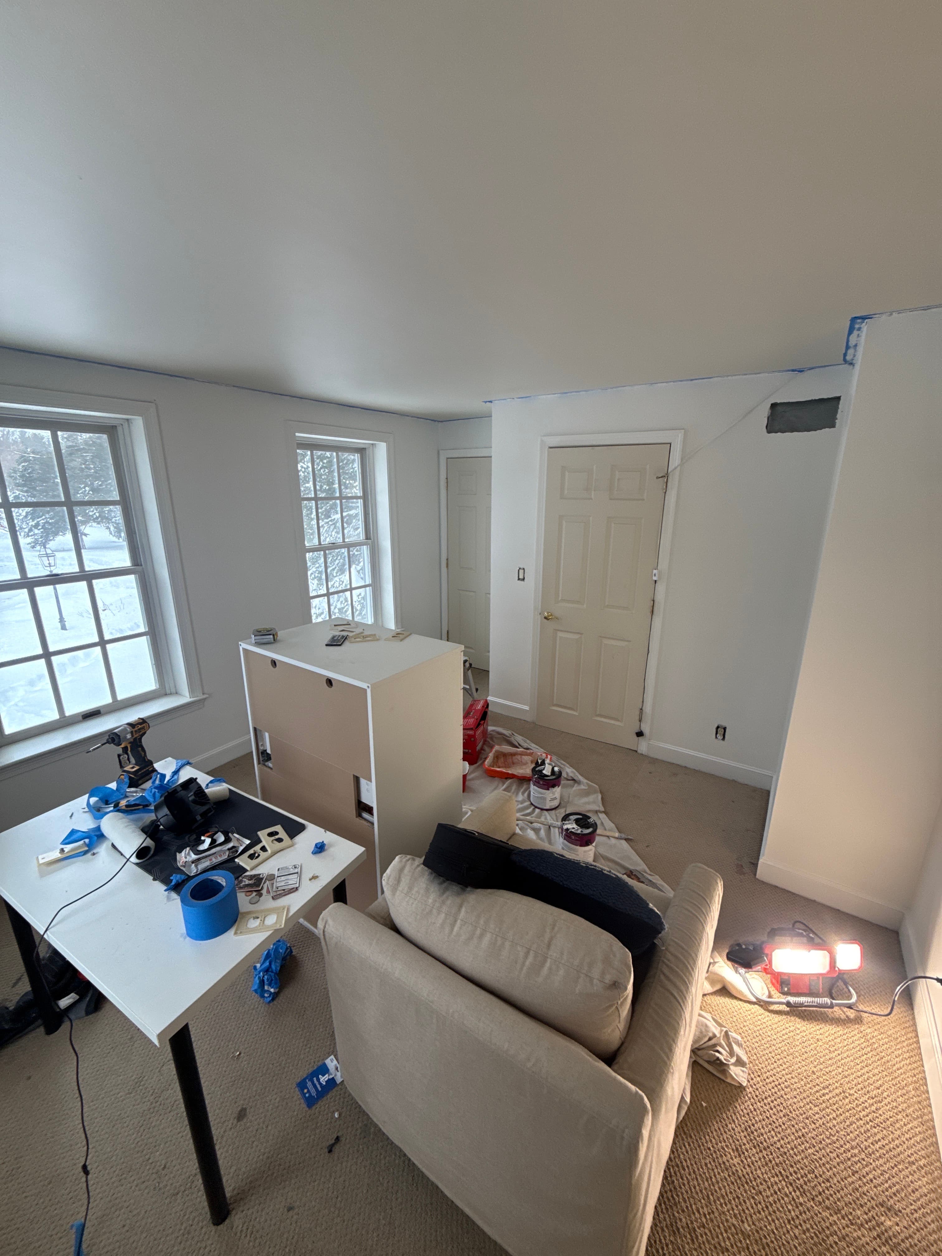 Room undergoing renovation with white walls, blue painter's tape, tools, and a beige armchair.