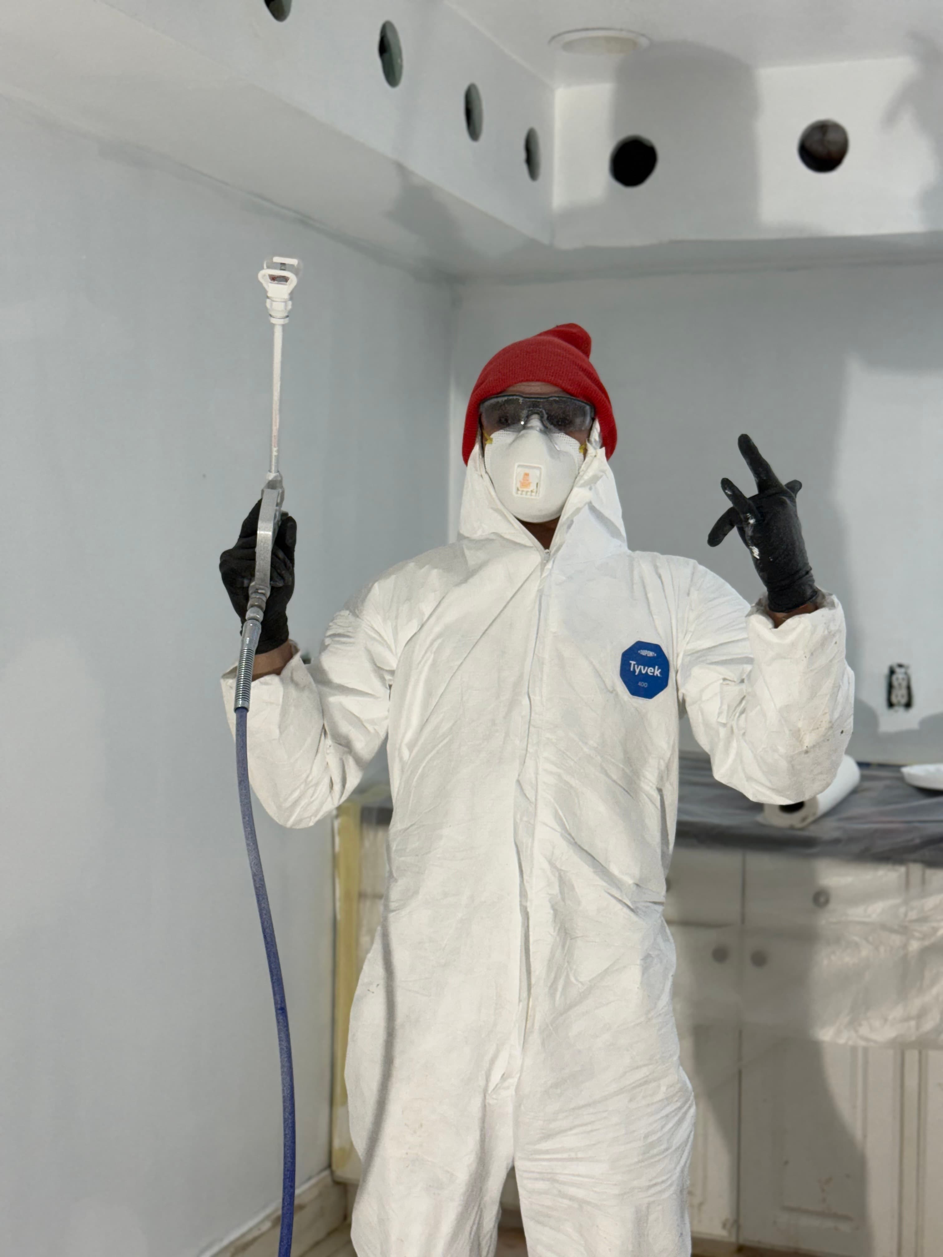 Painter in a white protective suit, red beanie, and respirator holding a paint sprayer wand.